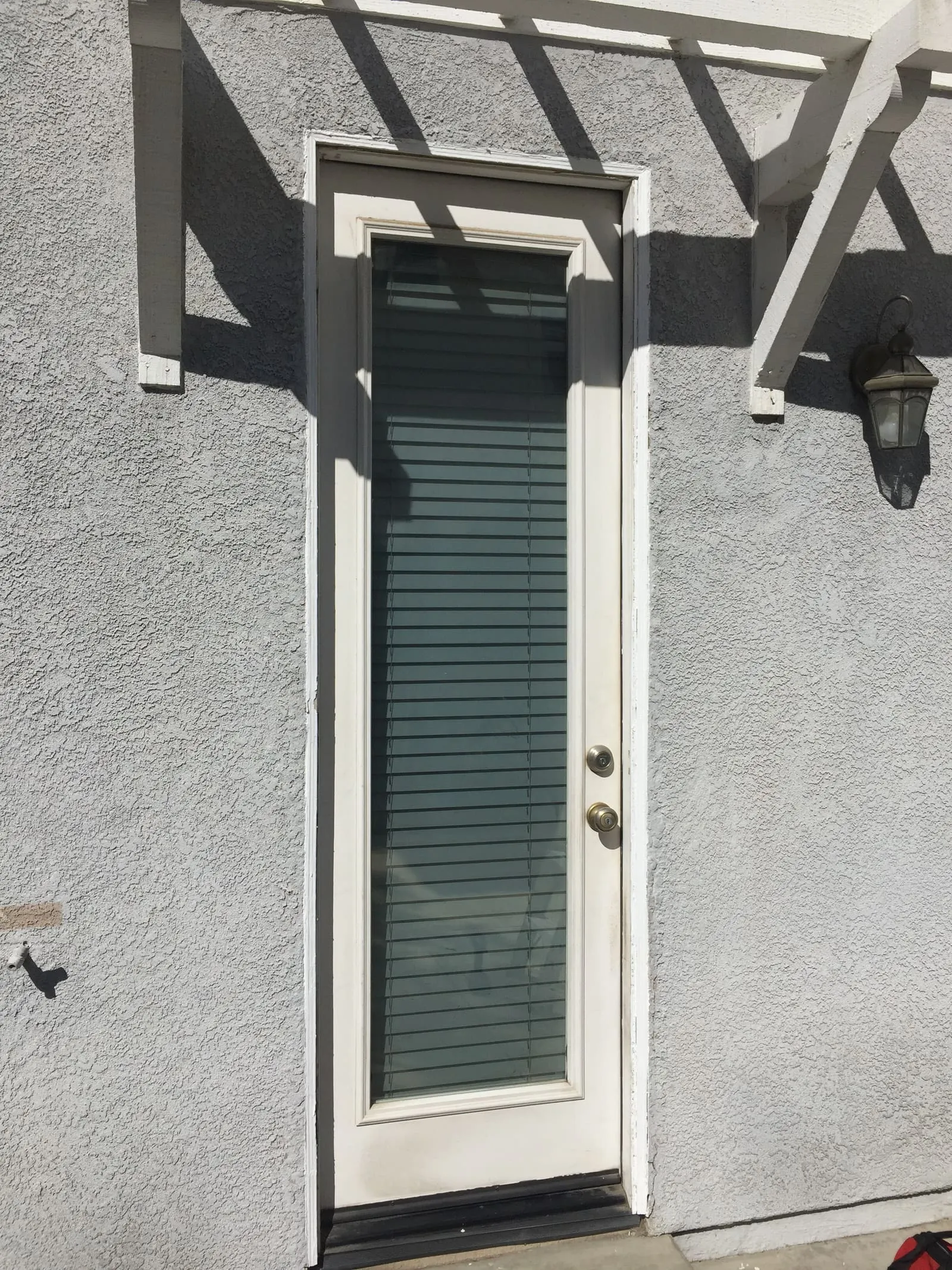 An entry door with vertical-blind glass and no screen, in bright sun on gray stucco