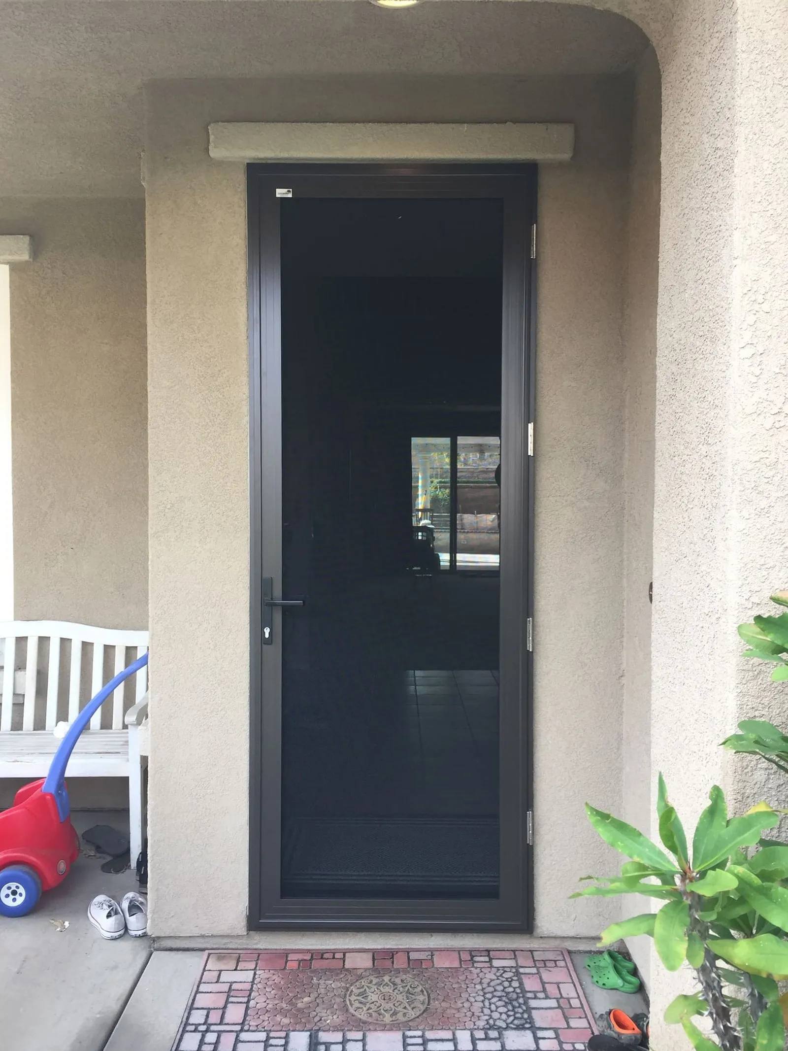Dark bronze hinged security screen door on a family porch with a welcome mat and toys