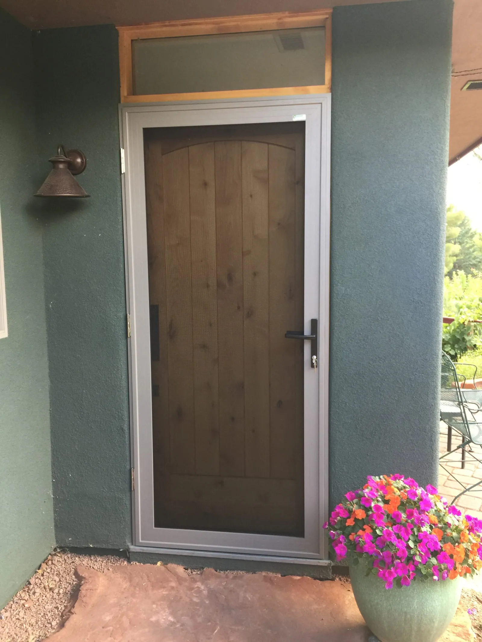 Gray-framed hinged security screen door on a green stucco entry with hanging flowers