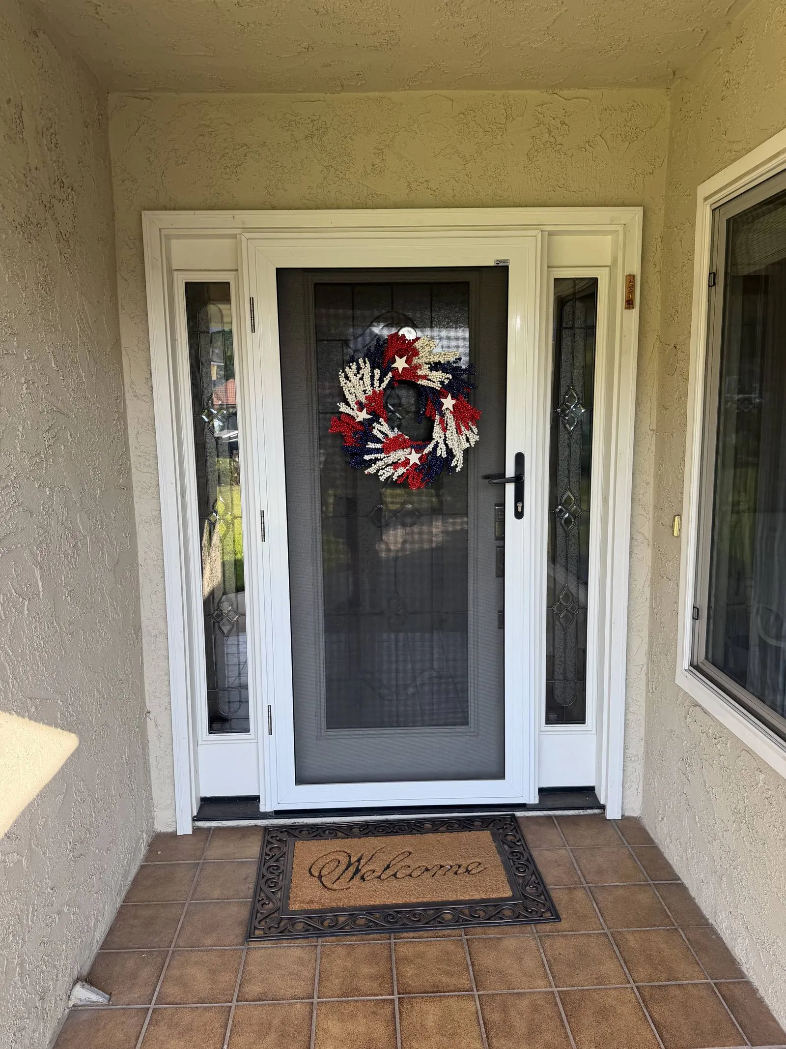 White full-view hinged security screen door on a stucco entry flanked by two leaded-glass sidelites, a red white and blue wreath hanging on the mesh